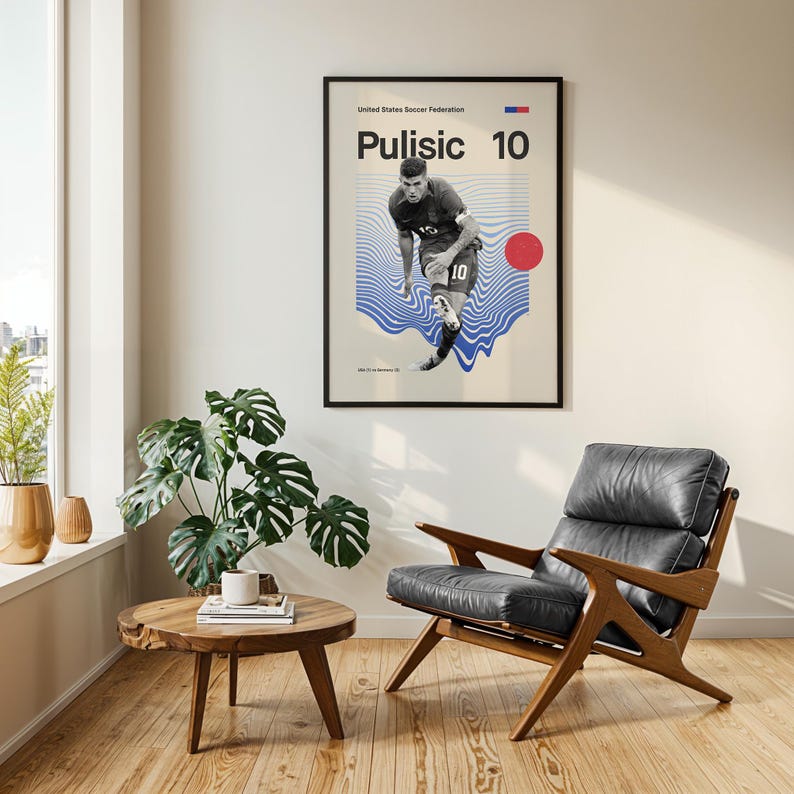Pu&ograve; includere: Un poster incorniciato con il calciatore Pulisic con il numero 10. Il poster ha uno sfondo beige con grafiche a onde blu e un cerchio rosso. La stanza comprende un tavolino in legno, una sedia in pelle e una pianta in vaso.