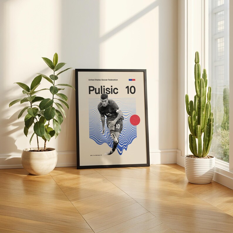 Pu&ograve; includere: Un poster incorniciato con il calciatore Pulisic, numero 10. Il poster ha uno sfondo beige con linee ondulate blu e un cerchio rosso. Il testo "United States Soccer Federation" &egrave; in alto.