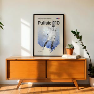 Pu&ograve; includere: Poster incorniciato con la scritta "Pulisic 10" e "United States Soccer Federation". Il poster presenta un calciatore e uno sfondo chiaro con motivi a onde blu. Il poster &egrave; in una cornice nera e viene visualizzato su un mobile in legno.