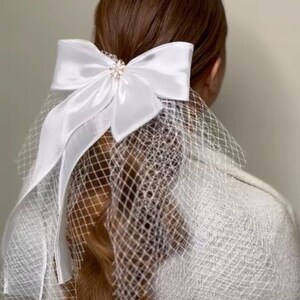 Puede incluir: Accesorio para el cabello de novia blanco con un gran lazo de satén y un pequeño adorno de piedras preciosas. El lazo está unido a un velo transparente.