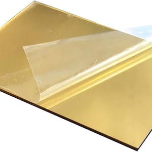 May include: A gold-coloured acrylic sheet with a protective film partially peeled away. The sheet has a reflective, mirror-like surface. The protective film is transparent with a blue tint. The sheet is rectangular.