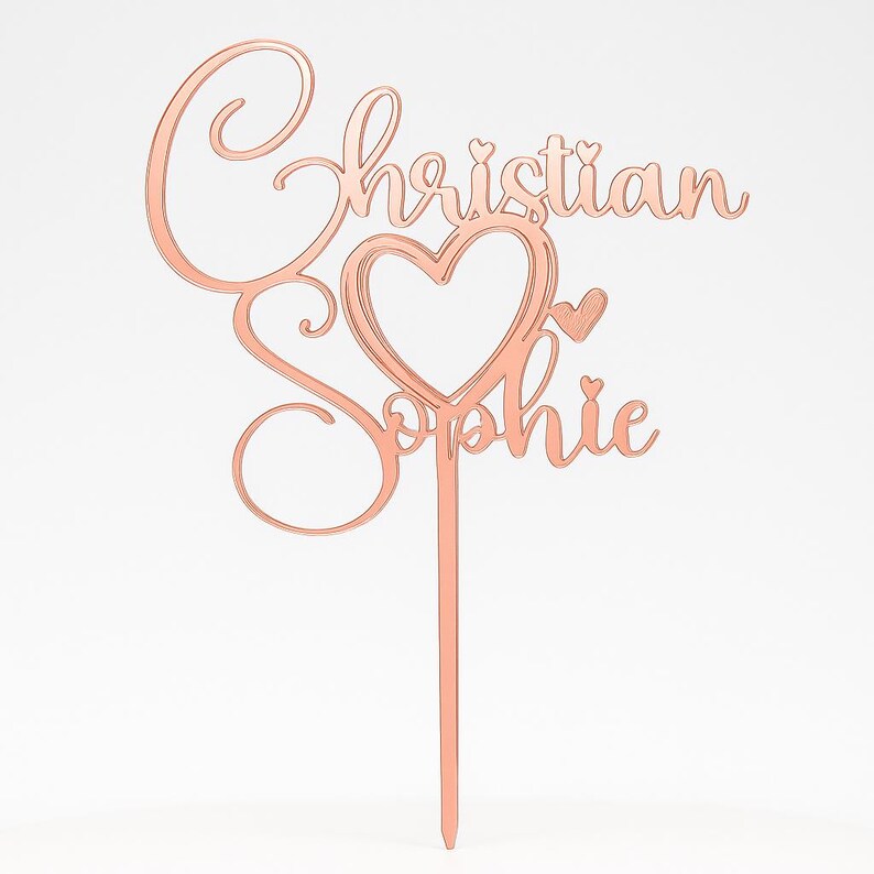 Peut inclure: D&eacute;coration de g&acirc;teau en or rose avec les noms "Christian" et "Sophie" en &eacute;criture cursive. Un c&oelig;ur est int&eacute;gr&eacute; au centre des noms. La d&eacute;coration est fix&eacute;e &agrave; une fine tige pointue pour &ecirc;tre ins&eacute;r&eacute;e dans un g&acirc;teau.