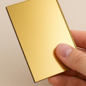 May include: A rectangular, gold-coloured object with a reflective surface. The object is held by a hand, showcasing its polished finish and metallic sheen. The edges are a darker shade of gold.