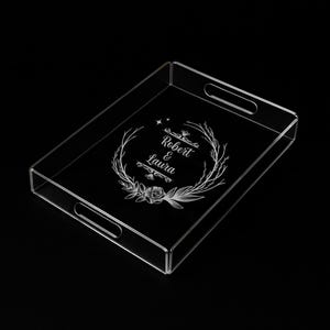 Peut inclure: Plateau de service en acrylique transparent avec poignées. Le plateau présente un motif de couronne florale gravée avec les noms "Robert & Laura" et une étoile. Le plateau est rectangulaire et de conception minimaliste.