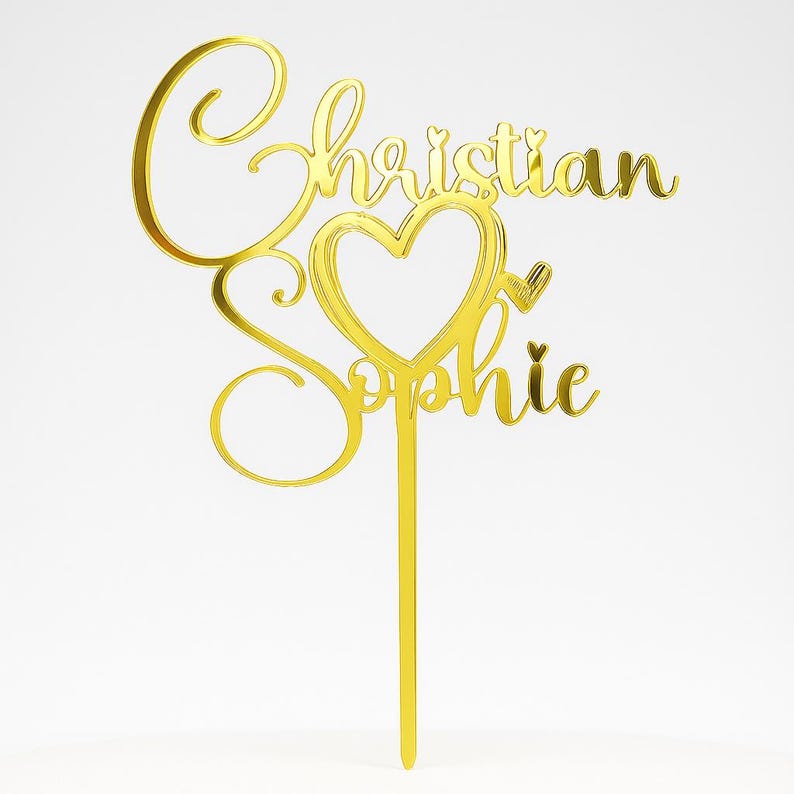 Peut inclure: Un cake topper dor&eacute; avec les noms "Christian" et "Sophie" &eacute;crits en cursive. Le design comprend un c&oelig;ur avec une fl&egrave;che et une tige pour l'insertion dans le g&acirc;teau. Le topper est fait d'un mat&eacute;riau r&eacute;fl&eacute;chissant.