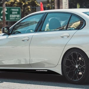 May include: A white sedan with black rims and a black side skirt featuring the word "PERFORMANCE" in white. The car is parked on a road, with a blurred background of trees and buildings.