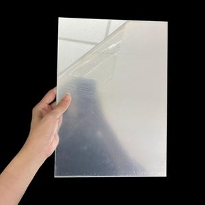 Puede incluir: Una lámina rectangular transparente de plástico con una película protectora parcialmente despegada. La lámina es sostenida por una mano sobre un fondo negro, mostrando su transparencia y superficie reflectante. El plástico parece liso y limpio.