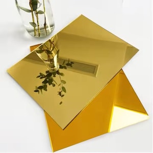 May include: Two gold-coloured, reflective acrylic sheets, one rectangular and one triangular, are stacked. A clear glass vase with green stems and leaves is reflected in the top sheet. The sheets are on a white surface.