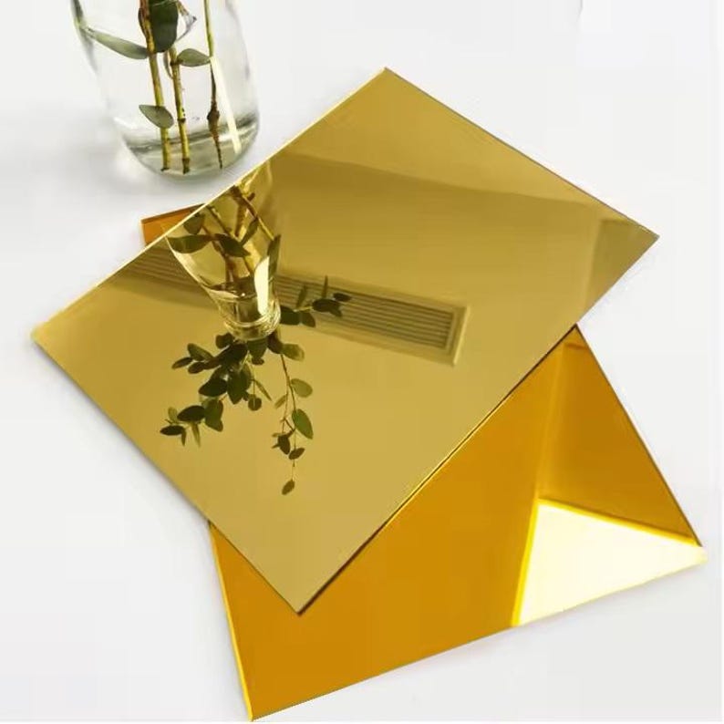 May include: Two gold-coloured, reflective acrylic sheets, one rectangular and one triangular, are stacked. A clear glass vase with green stems and leaves is reflected in the top sheet. The sheets are on a white surface.