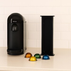 Op de afbeelding: Een zwarte Vertuo koffiemachine met een bijpassende capsulehouder. De machine heeft een afgeronde bovenkant en een strak design. Verschillende kleurrijke koffiecapsules in goud, groen, geel en blauw liggen voor de machine.