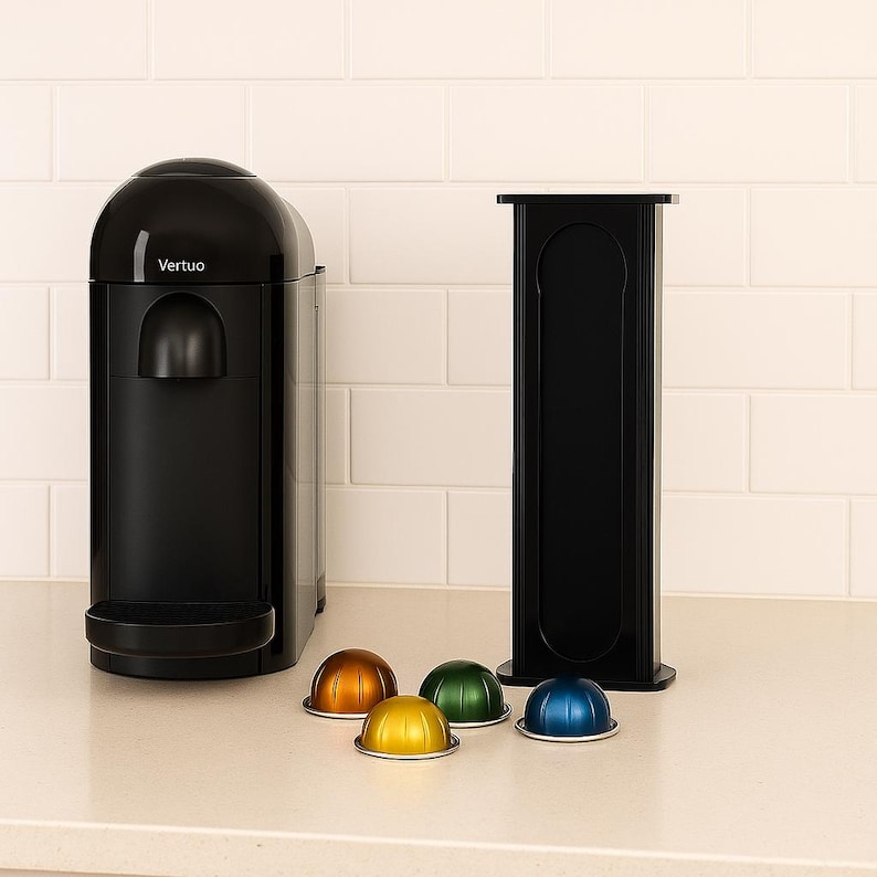 Op de afbeelding: Een zwarte Vertuo koffiemachine met een bijpassende capsulehouder. De machine heeft een afgeronde bovenkant en een strak design. Verschillende kleurrijke koffiecapsules in goud, groen, geel en blauw liggen voor de machine.
