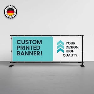 Op de afbeelding: Een op maat bedrukte banner met de tekst "CUSTOM PRINTED BANNER!" in grote, vette letters. De banner bevat ook de tekst "YOUR DESIGN, HIGH QUALITY." De banner wordt ondersteund door een zwart metalen frame.