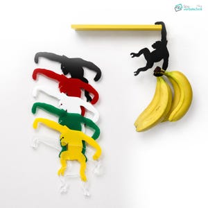 May include: A set of colourful monkey-shaped wall hooks in black, red, white, green, and yellow. A yellow shelf is above the hooks, and a black monkey hook holds two bananas. The design is modern and playful.