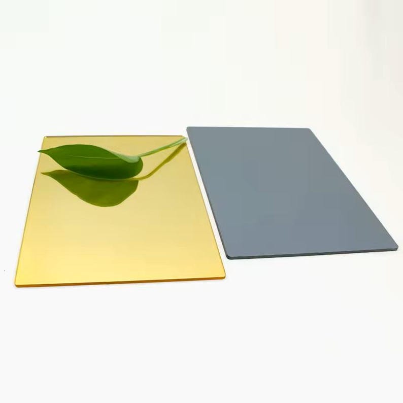 May include: Two rectangular sheets, one gold and one grey, are displayed against a white background. A green leaf rests on the gold sheet. The gold sheet has a reflective, mirror-like surface. The grey sheet has a matte finish.