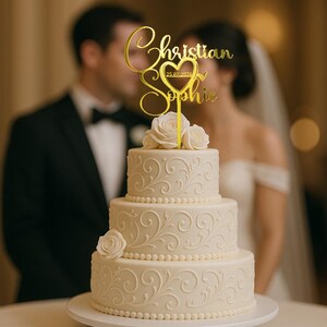 Peut inclure: Un g&acirc;teau de mariage &agrave; trois &eacute;tages avec un gla&ccedil;age blanc et des motifs floraux. Un cake topper dor&eacute; indique "Christian & Sophie" avec un c&oelig;ur et une date. Des roses blanches ornent le premier &eacute;tage. Un couple est flou en arri&egrave;re-plan.