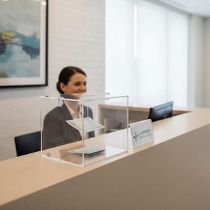 May include: A clear acrylic suggestion box with a slot on top, sitting on a light wood counter. The box contains papers. A sign that says "SUPPORT PATIENT CARE" is on the counter. A person is visible behind the box.