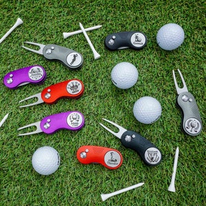 Personalisierbares Golftool von WOLFSARTIG, bestehend aus Pitchgabel und Golfballmarker, verfügbar in den Farben Schwarz, Wildberry, Rubinrot und Anthrazit. Fotografiert auf einem Golfplatz, umgeben von Golfbällen und Tees.