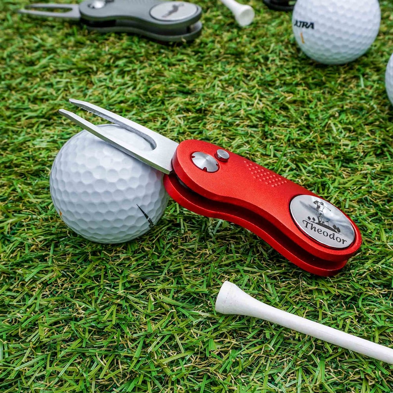 Ein rubinrotes Golftool von WOLFSARTIG, angelehnt an einen Golfball auf einer Wiese. Der Golfballmarker ist mit einem Namen und dem Motiv eines Golfspielers durch Lasergravur personalisiert.