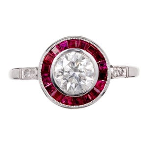 Puede incluir: Un anillo de platino con un diamante central claro y redondo, rodeado por un halo de piedras preciosas rojas oscuras rectangulares. La banda tiene pequeñas piedras claras a cada lado.