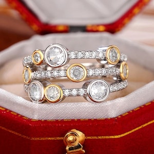 May include: A multi-layered ring with a silver band and gold accents. The ring features several large, clear, round stones set in gold bezels, interspersed with smaller, clear stones. The ring is displayed in a red jewelry box.