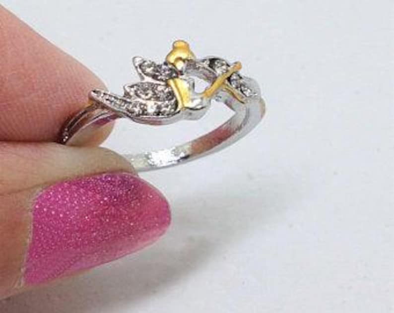 K&ouml;nnte beinhalten: Ein silberner Ring mit einem goldfarbenen Design eines Vogels mit Fl&uuml;geln und einer Krone auf dem Kopf. Der Vogel ist mit klaren Strasssteinen verziert.