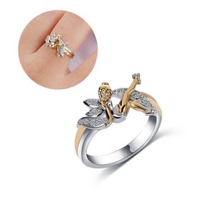 K&ouml;nnte beinhalten: Ein silberner Ring mit einem goldenen Band und einem Feendesign mit klaren Steinen. Die Fee sitzt auf einem Ast mit ausgebreiteten Fl&uuml;geln.