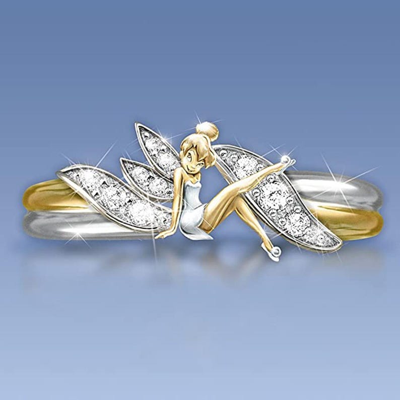 K&ouml;nnte beinhalten: Ein goldener und silberner Ring mit einem Feendesign. Die Fee sitzt auf dem Ring mit ausgebreiteten Fl&uuml;geln. Der Ring ist mit kleinen, klaren Steinen verziert.