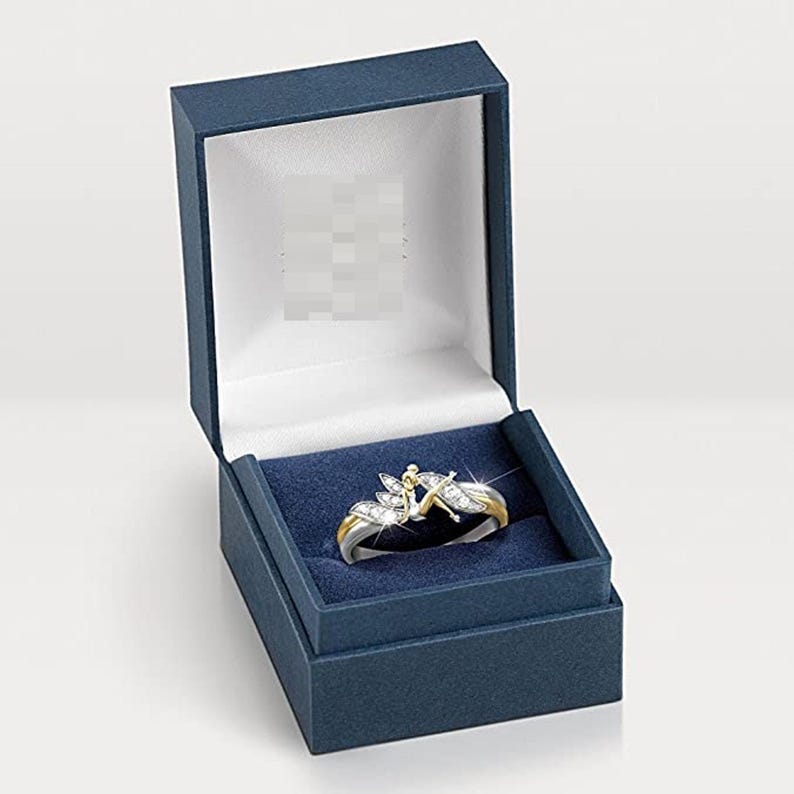 K&ouml;nnte beinhalten: Ein silberner Ring mit goldener Verzierung und einem klaren Kristalldesign mit einer Fee. Der Ring wird in einer blauen Samt-Ringbox pr&auml;sentiert.