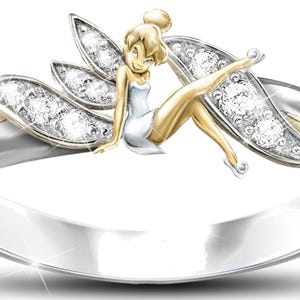 K&ouml;nnte beinhalten: Ein silberner Ring mit einer goldenen Feenfigur, die darauf sitzt. Die Fee hat Fl&uuml;gel mit klaren Steinen und tr&auml;gt ein wei&szlig;es Kleid. Der Ring ist mit klaren Steinen besetzt.