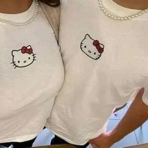 May include: Two white t-shirts with a Hello Kitty graphic on the front. The shirts are worn by two people. The person on the left is wearing a pearl necklace.