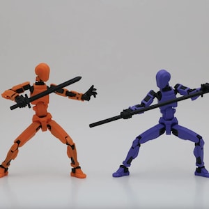 Puede incluir: Dos figuras de acción articuladas en poses dinámicas. Una es naranja y empuña una espada negra, la otra es morada con un bastón negro. Ambas figuras están sobre un fondo blanco liso, lo que sugiere un tema de artes marciales o acción.