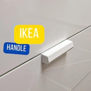 May include: A white cabinet handle with a rectangular shape. The handle is mounted on a white cabinet door. The image also includes the text "IKEA HANDLE" in a yellow and blue design.