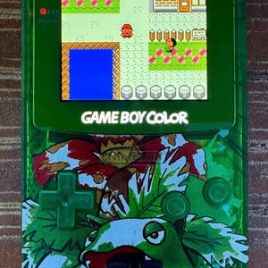 Puede incluir: Consola de juegos portátil Game Boy Color verde con un diseño de Pokemon. La consola presenta una imagen verde y blanca de un personaje Pokemon con una boca grande y una lengua roja. La pantalla muestra una escena de juego pixelada con un personaje rojo y una masa de agua azul.