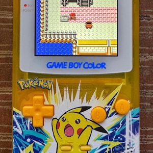 Puede incluir: Una consola de videojuegos portátil Game Boy Color amarilla con un diseño de Pikachu. La pantalla muestra un juego de Pokemon. La consola tiene un esquema de color amarillo, azul y blanco. Los botones están etiquetados como A, B, Select y Start.