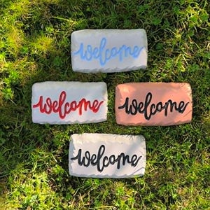 May include: Four rectangular stone-like plaques with textured edges, each featuring the word welcome in cursive script. One plaque is white with light blue text, one is white with red text, one is peach with black text, and one is white with black text.