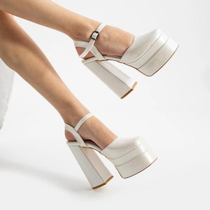 May include: White platform sandals with thick heels and ankle straps. The sandals have a stacked platform sole and a square toe. The heels are a chunky, rectangular shape. The sandals are made of a smooth, shiny material.