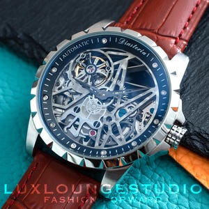 May include: An automatic wristwatch with a silver-tone stainless steel case and a skeletonized dial revealing internal mechanical components. The watch features a dark blue bezel with white markers and a brown leather strap with a crocodile texture.