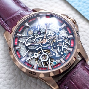 Könnte beinhalten: Eine roségoldene Skelettuhr mit einem burgunderfarbenen Lederarmband. Das Zifferblatt der Uhr hat eine klare Glasabdeckung und rote Akzente am Rand.