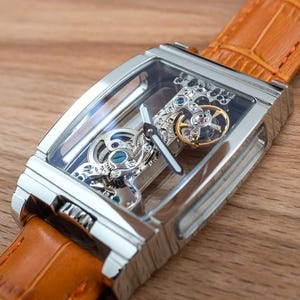 May include: A rectangular, silver-toned skeleton watch with a transparent face, showcasing the intricate internal mechanisms. The watch features a gold-colored gear and is attached to a tan leather strap. The watch is displayed on a wooden surface.