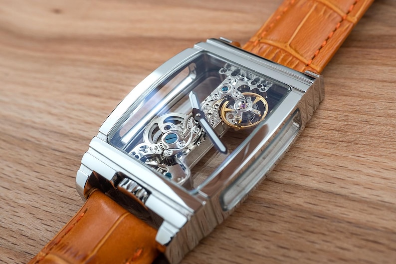 May include: A rectangular, silver-toned skeleton watch with a transparent face, showcasing the intricate internal mechanisms. The watch features a gold-colored gear and is attached to a tan leather strap. The watch is displayed on a wooden surface.