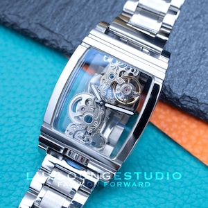 Puede incluir: Un reloj esqueleto plateado con una esfera transparente y una correa de metal plateado. La esfera del reloj muestra el intrincado funcionamiento interno del reloj.