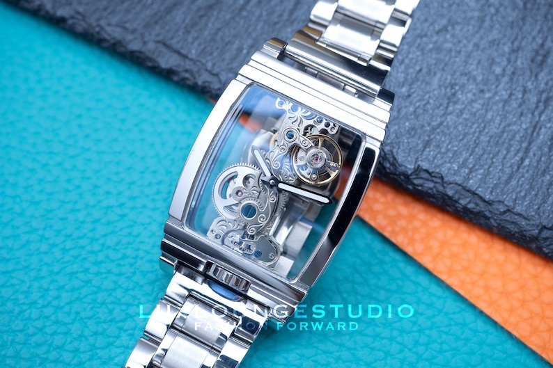 Puede incluir: Un reloj esqueleto plateado con una esfera transparente y una correa de metal plateado. La esfera del reloj muestra el intrincado funcionamiento interno del reloj.