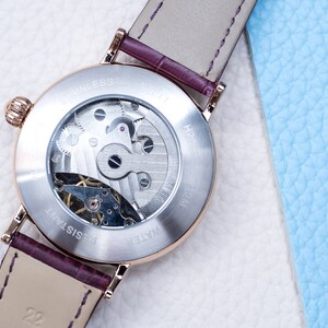 May include: A silver watch with a brown leather strap. The back of the watch is clear, revealing the inner workings of the watch. The watch face has the text "WATER RESISTANT" and "STAINLESS STEEL" printed on it.