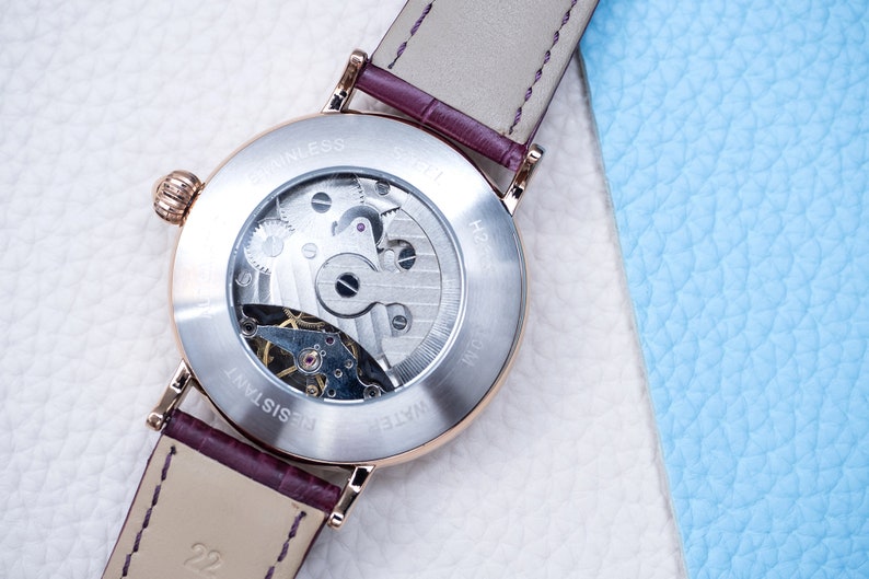 May include: A silver watch with a brown leather strap. The back of the watch is clear, revealing the inner workings of the watch. The watch face has the text "WATER RESISTANT" and "STAINLESS STEEL" printed on it.