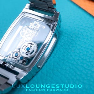 Puede incluir: Un reloj plateado con una parte posterior transparente que revela el intrincado funcionamiento mec&aacute;nico. El reloj tiene una correa plateada y una esfera rectangular. La esfera del reloj tiene la palabra "Diasterna" impresa. El reloj est&aacute; sobre una superficie turquesa.