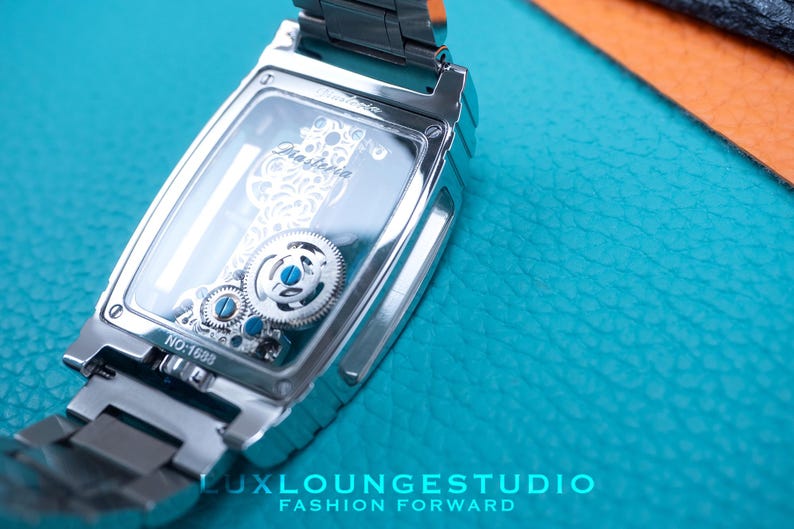 Puede incluir: Un reloj plateado con una parte posterior transparente que revela el intrincado funcionamiento mec&aacute;nico. El reloj tiene una correa plateada y una esfera rectangular. La esfera del reloj tiene la palabra "Diasterna" impresa. El reloj est&aacute; sobre una superficie turquesa.