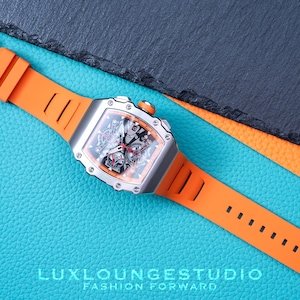 Peut inclure: Une montre de luxe avec un bo&icirc;tier argent&eacute; et un bracelet en silicone orange. Le cadran de la montre est noir avec des d&eacute;tails complexes. Le bracelet est fix&eacute; avec une boucle argent&eacute;e. La montre est pr&eacute;sent&eacute;e sur une surface bleue textur&eacute;e.