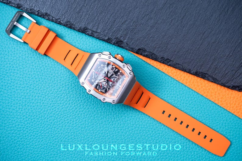 Peut inclure: Une montre de luxe avec un bo&icirc;tier argent&eacute; et un bracelet en silicone orange. Le cadran de la montre est noir avec des d&eacute;tails complexes. Le bracelet est fix&eacute; avec une boucle argent&eacute;e. La montre est pr&eacute;sent&eacute;e sur une surface bleue textur&eacute;e.