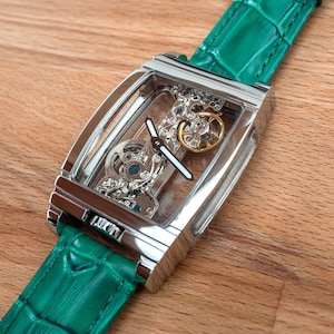 May include: A rectangular, silver skeleton watch with a green leather strap. The watch face displays intricate internal mechanisms, including silver and gold-coloured gears. The strap is a vibrant green, and the watch is displayed on a wooden surface.