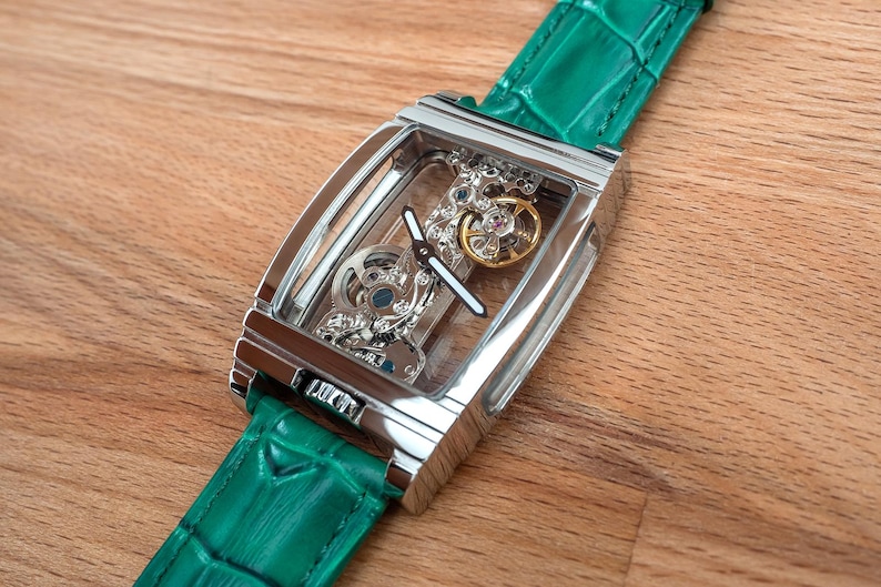 May include: A rectangular, silver skeleton watch with a green leather strap. The watch face displays intricate internal mechanisms, including silver and gold-coloured gears. The strap is a vibrant green, and the watch is displayed on a wooden surface.
